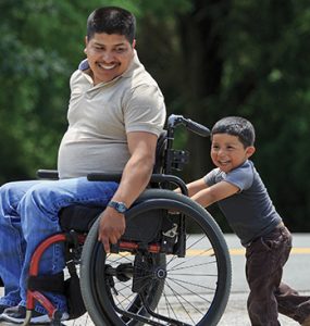Young father in a wheelchair being pushed by his young son.
