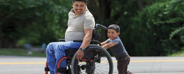 Young father in a wheelchair being pushed by his young son.