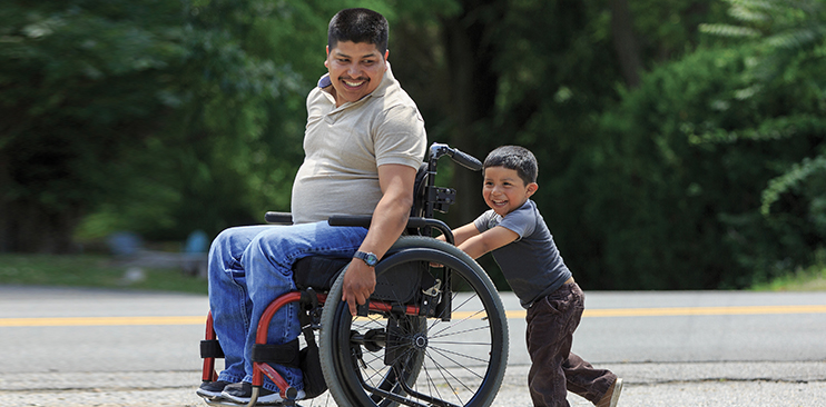 Young father in a wheelchair being pushed by his young son.