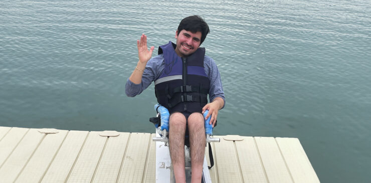 A young man sit on his wakeboard on a dock overlooking a large ontario lake.